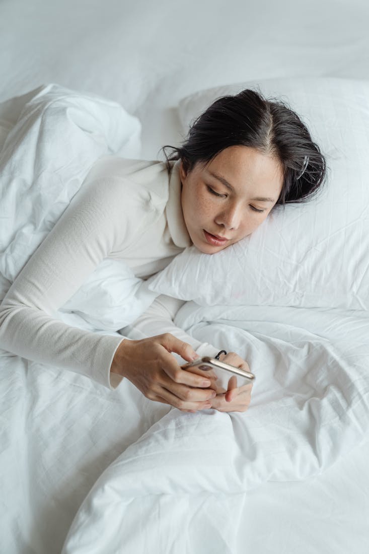 From above calm young Asian female in sleepwear waking up in morning and browsing mobile phone in cozy bed